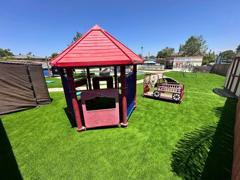 Artificial grass preschool play yard in San Diego with a red-roofed playhouse and a toy train under a clear blue sky, installed by Turbo Surfacing Solutions