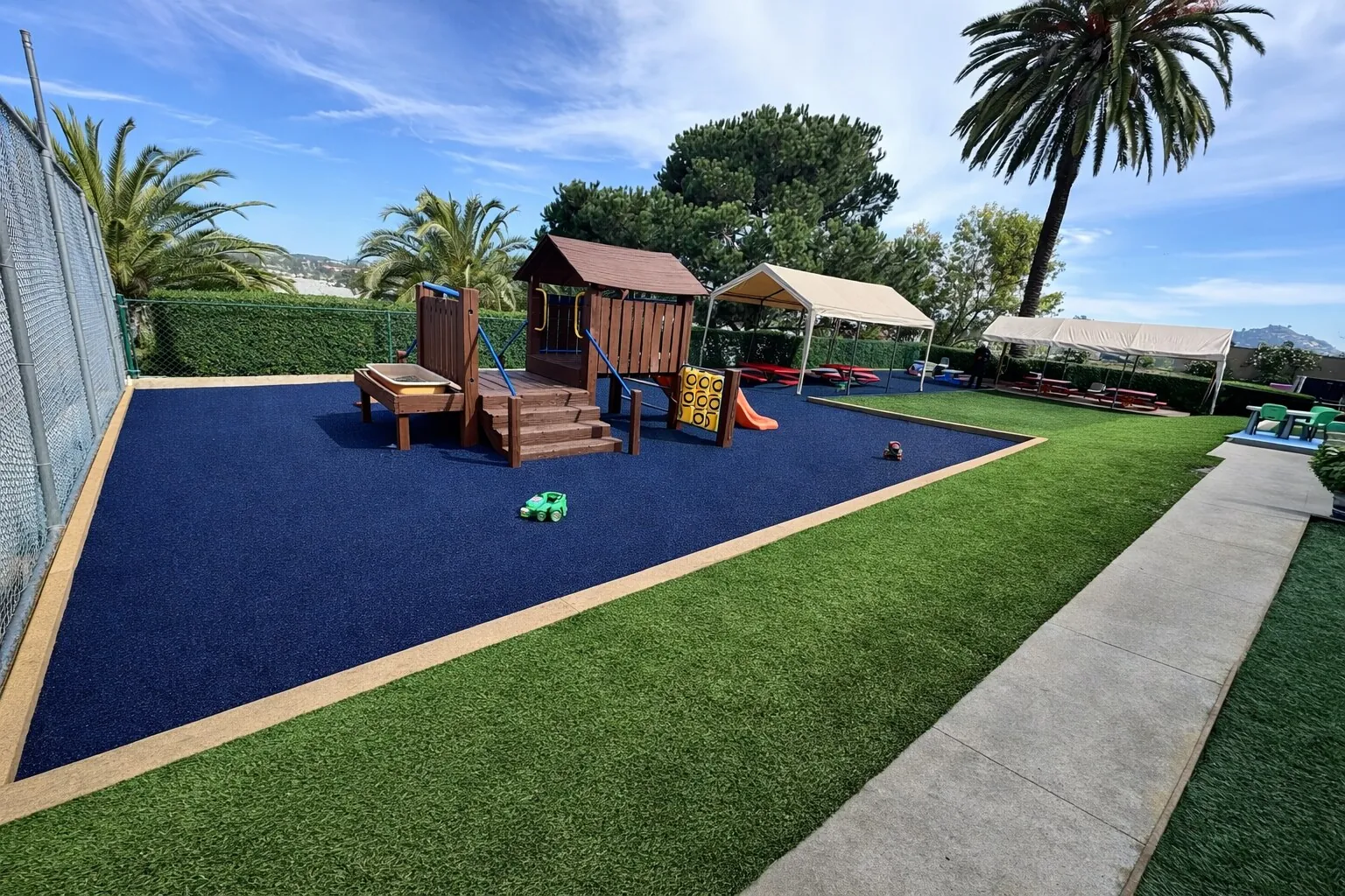 Navy blue poured-in-place rubber playground surface with wooden play structure, artificial grass and palm trees at a sunny San Diego County preschool